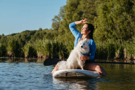 Happy Young Woman on the Lake at Early Morning Sitting on the Sup Board with Her Dog Snow-White Japanese Spitz and Enjoying Sunriseの写真素材