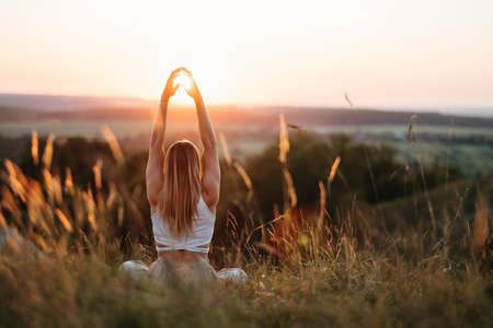 Back View on Woman Sitting in Meditation Yoga Pose and Catching Sun by Hands at Sunset Outdoorsの写真素材