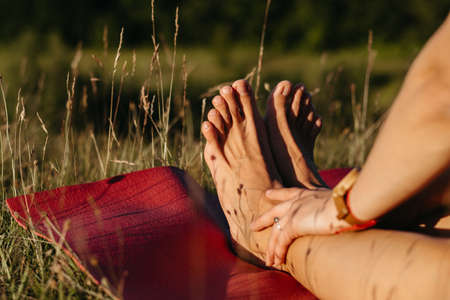 Close Up Unrecognisable Woman Holding Her Feet While Sitting on the Mat During Yoga Outdoors at Sunsetの写真素材