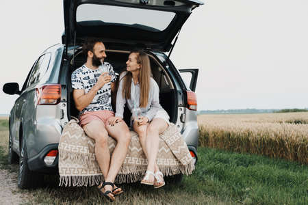 Caucasian cheerful woman and man drinking coffee in trunk of the car, middle-aged couple making stop while on a road tripの写真素材