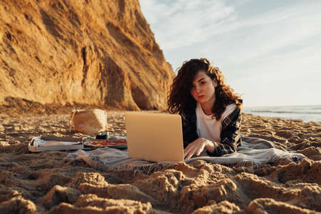 Young curly haired woman working on laptop while laying on the seashore at dawnの写真素材