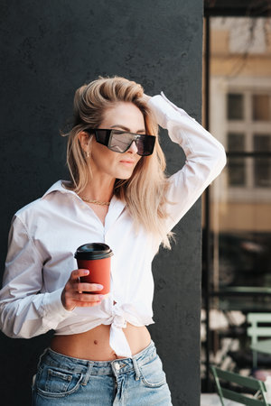 Elegant Blonde Woman Holding Cup of Coffee in Hand and Posing near the Wall on Terrace of Cafe, Confident and Fashionable Female Model Wearing Sunglasses and White Shirt.の写真素材