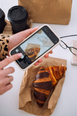 Woman taking photos of her lunch on smartphone, delicious croissants and cups of coffee on the...