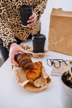 Woman at the workplace has a lunch of brown and chocolate croissants with coffee, food delivery conceptの写真素材