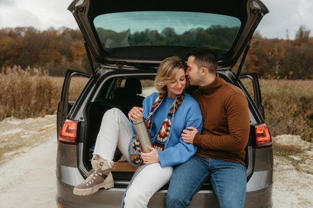 Happy adult couple sitting in the open trunk of a car while traveling in autumn, road trip concept,...
