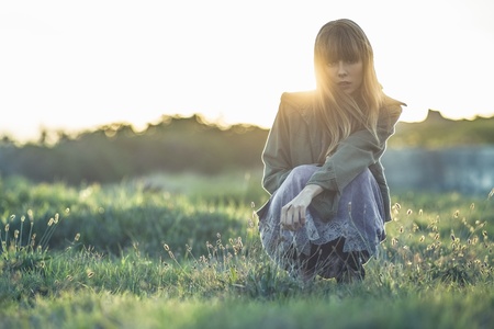 Fashionable young girl crouching in sheer dress and jacket staring at camera in a meadowの写真素材