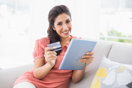 Smiling brunette sitting on her sofa using tablet to shop online at home in the living roomの写真素材