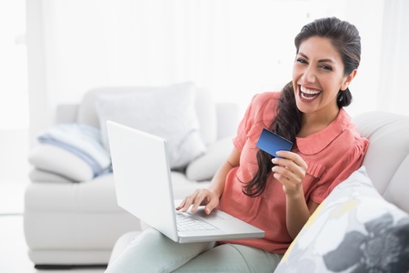 Laughing brunette sitting on her sofa using laptop to shop online at home in the sitting roomの写真素材