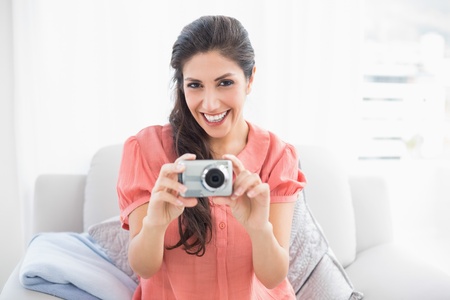 Happy brunette sitting on her sofa taking a picture of the camera at home in the sitting roomの写真素材