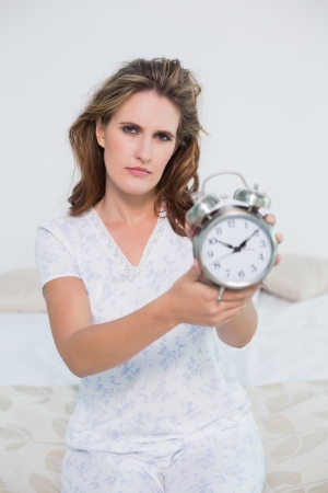 Frowning tired woman in bright bedroom showing alarm clockの写真素材