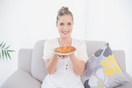 Cheerful fresh model holding croissant sitting on sofa in bright living roomの写真素材