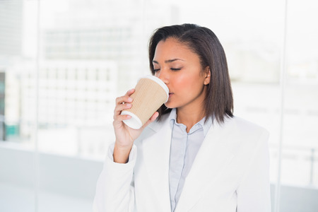 Cute young dark haired businesswoman enjoying coffee in bright officeの写真素材