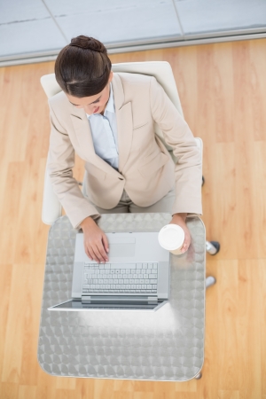 Attractive smart brown haired businesswoman using a laptop while holding a cup of coffee in bright officeの写真素材
