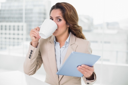 Businesswoman using tablet drinking coffee looking at camera in bright officeの写真素材