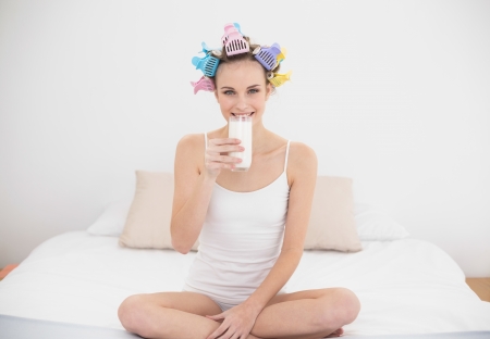 Attractive natural brown haired woman in hair curlers drinking milk in bright bedroomの写真素材