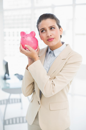 Frowning smart brown haired businesswoman shaking a piggy bank in bright officeの写真素材
