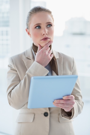 Blonde pensive businesswoman holding tablet in bright officeの写真素材