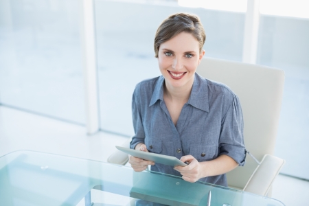 Cheerful businesswoman holding her tablet sitting at her desk smiling at cameraの写真素材