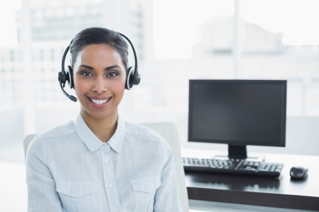 Attractive female agent wearing headset smiling at camera sitting in bright officeの写真素材