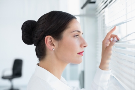 Close-up side view of a young business woman peeking through blinds at officeの写真素材