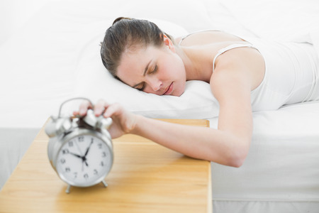 Sleepy woman in bed extending hand to blurred alarm clock at bedroomの写真素材