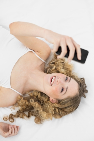 Pretty smiling blonde lying on bed holding mobile phone in bright bedroomの写真素材