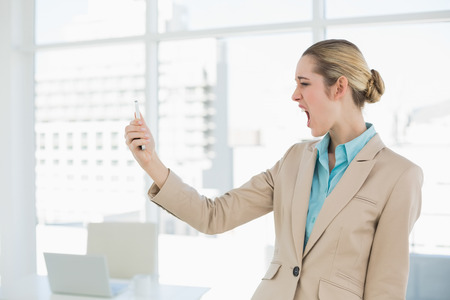 Attractive young businesswoman looking shocked at her smartphone standing in her officeの写真素材