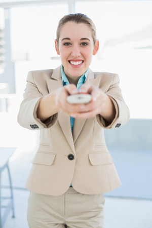 Attractive businesswoman holding a remote smiling cheerfully at camera standing in her officeの写真素材