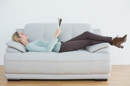 Attractive blonde woman reading newspaper lying on couch in living roomの写真素材