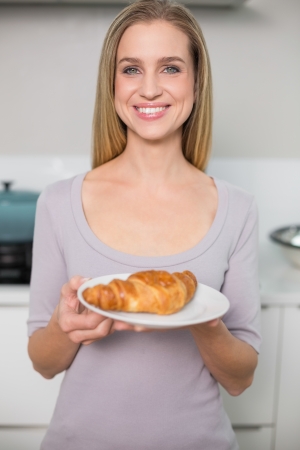 Cheerful gorgeous model holding plate with croissant standing in kitchenの写真素材