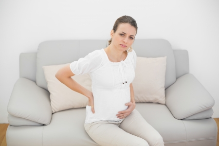 Suffering pregnant woman looking seriously at camera touching her injured back sitting on couchの写真素材
