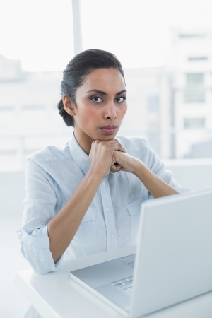 Serious dark haired woman looking at camera sitting at her desk in bright officeの写真素材