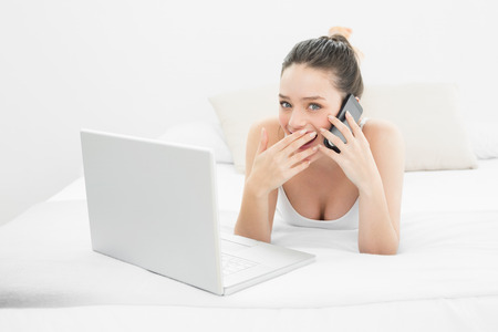 Portrait of a smiling casual young woman using cellphone and laptop in bedの写真素材