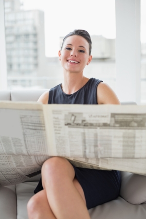 Portrait of a smiling beautiful businesswoman reading newspaper on sofa at a bright homeの写真素材