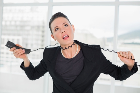 Frustrated young elegant businesswoman with telephone cable around her neck at a bright officeの写真素材