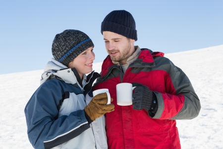 Smiling young couple in warm clothing with coffee cups standing on snow covered landscapeの写真素材