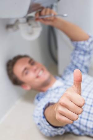 Portrait of a young plumber repairing washbasin drain while gesturing thumbs up in bathroomの写真素材