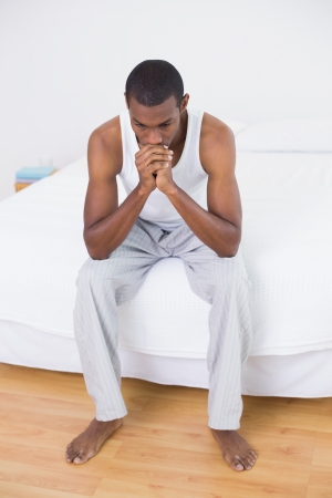 Full length of a thoughtful young Afro man sitting on bed at homeの写真素材