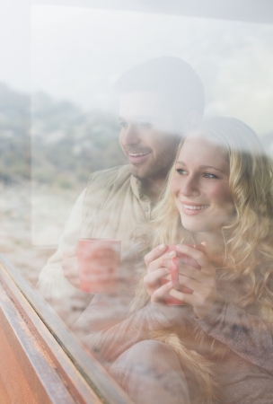 Thoughtful happy young couple with coffee cups looking out through cabin windowの写真素材