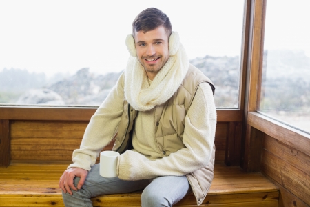Portrait of a smiling young man wearing earmuff with coffee cup against cabin windowの写真素材