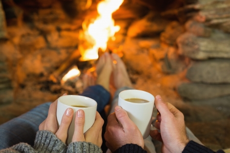 Close up of hands holding coffee cups in front of lit fireplaceの写真素材
