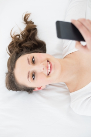 High angle portrait of a young brunette with mobile phone in bed at homeの写真素材