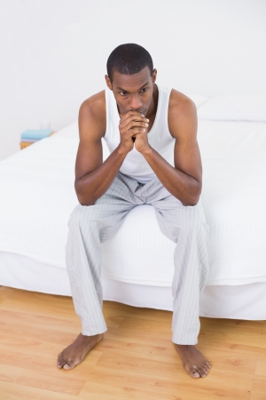 Full length of a thoughtful young Afro man sitting on bed at homeの写真素材