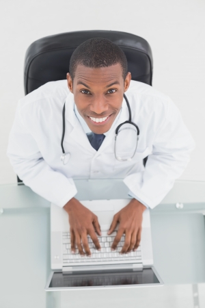 Overhead portrait of a smiling male doctor using laptop at medical officeの写真素材