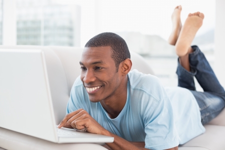 Relaxed casual young Afro man with laptop lying on sofa in a bright houseの写真素材