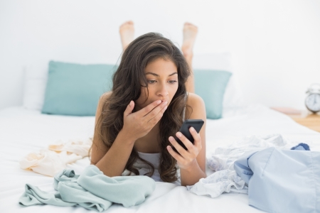 Shocked young woman looking at mobile phone in bed at homeの写真素材