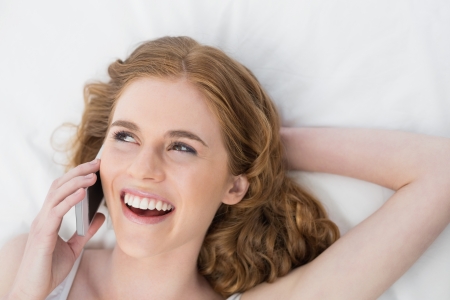 Cheerful young woman using mobile phone in bed at homeの写真素材