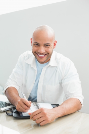 Portrait of a casual smiling young man with laptop writing in diary at homeの写真素材