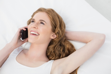 Cheerful young woman using mobile phone in bed at homeの写真素材