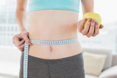 Close up mid section of a young woman measuring waist as she holds apple in fitness studioの写真素材